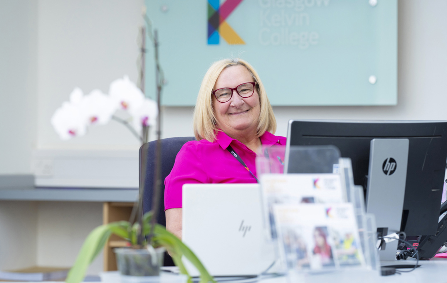 Receptionist smiling at her desk in the Glasgow Kelvin College West Campus, with a Glasgow Kelvin College sign in the background and orchids in the foreground.