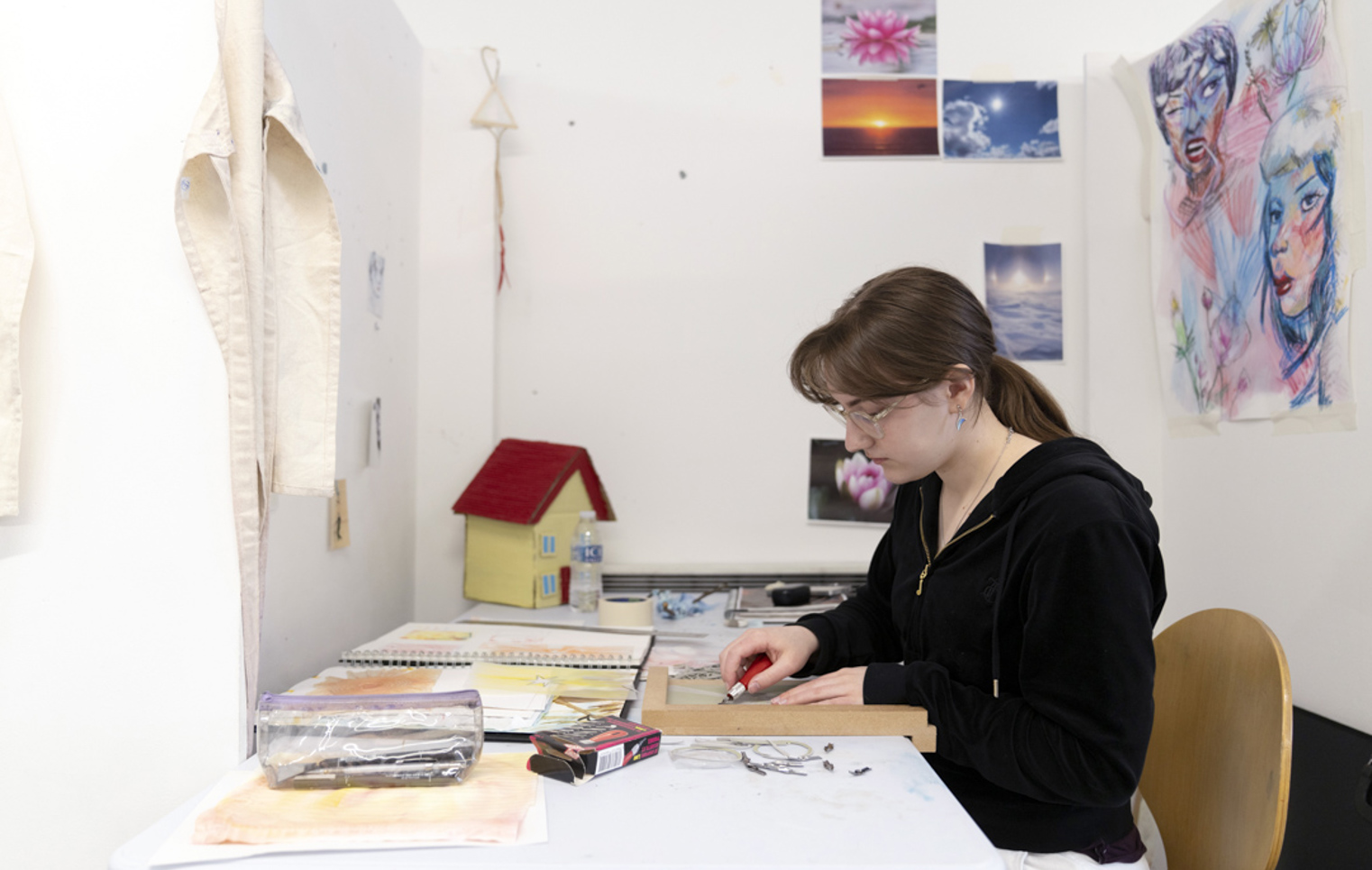 Person seated at a desk in an art studio, carving a design into a printmaking block, with sketchbooks and artwork on the wall