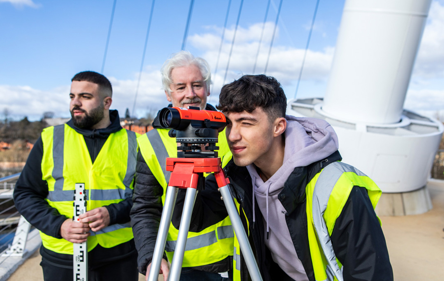 A young student in a high-visibility vest looking through a surveying instrument while the lecturer and another student observe and discuss measurements on a bridge