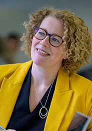 Maria Bradshaw, a woman with curly blonde hair and glasses, wearing a bright yellow blazer and black top, smiling in a professional setting.