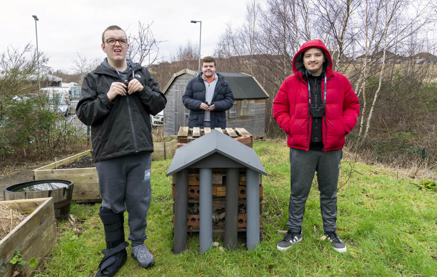 Three young men standing outside beside a handmade bug hotel, surrounded by greenery and raised garden beds.