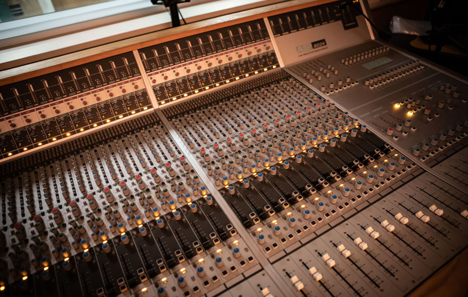 A wide shot of a professional recording studio mixing desk, with multiple sliders, knobs, and glowing lights.
