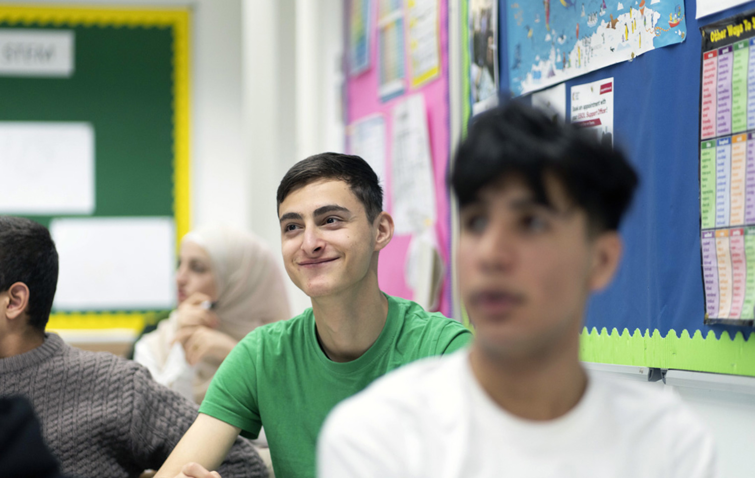 A young male student in a green t-shirt sits in a classroom, smiling while holding a whiteboard marker. Other students are visible in the background, including a student in a white hijab.