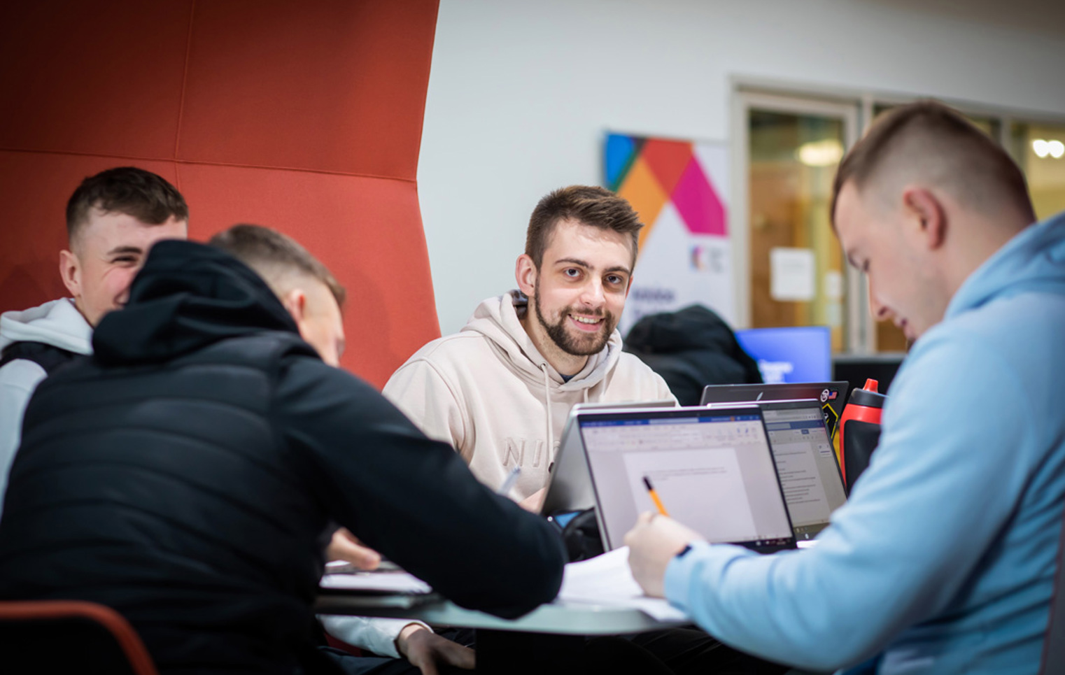 Group of male students working together on laptops and papers in a study pod at Glasgow Kelvin College’s Springburn Campus, with one student smiling at the camera.