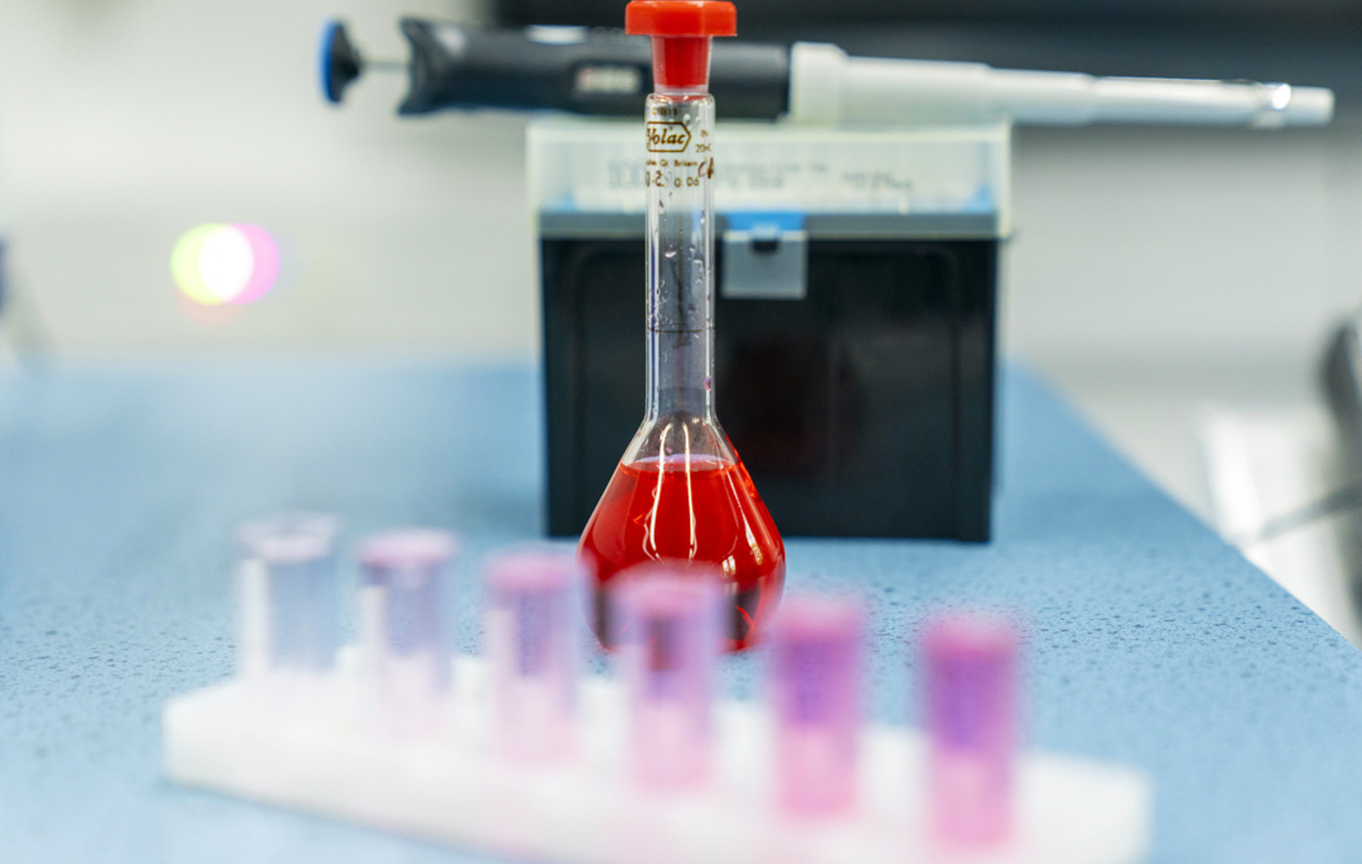 A bright red liquid fills a glass flask on a blue lab bench, with a row of test tubes in soft pinks and purples in front and a pipette resting in the background—science in action and full of colour.