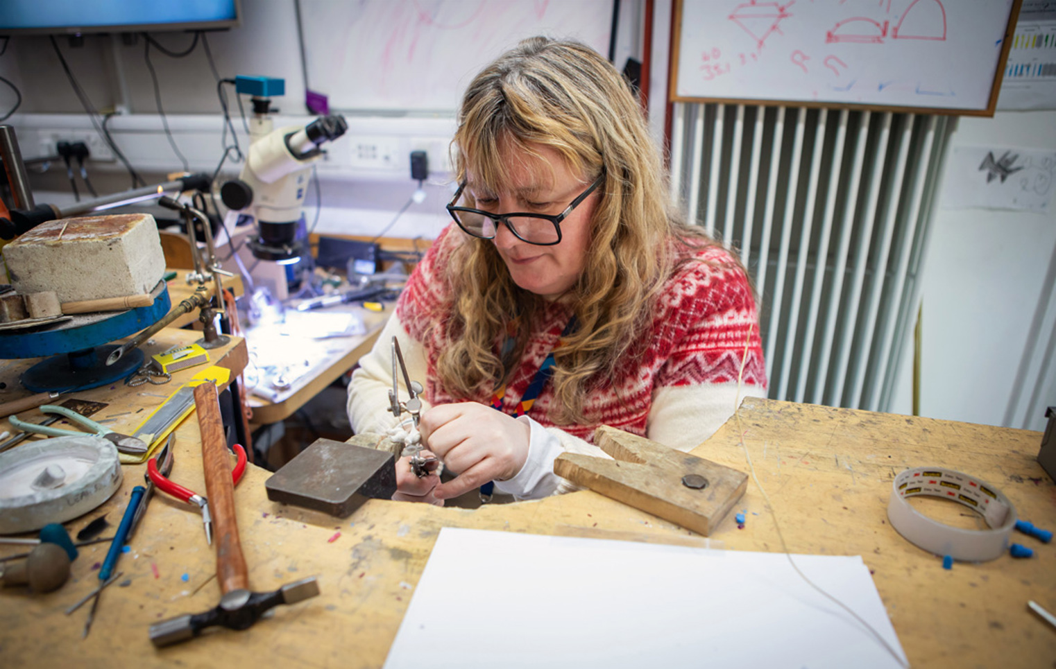 A female jewellery lecturer with long blonde hair and glasses, wearing a red and white sweater, working at a jewellery-making bench in a workshop filled with tools and equipment.