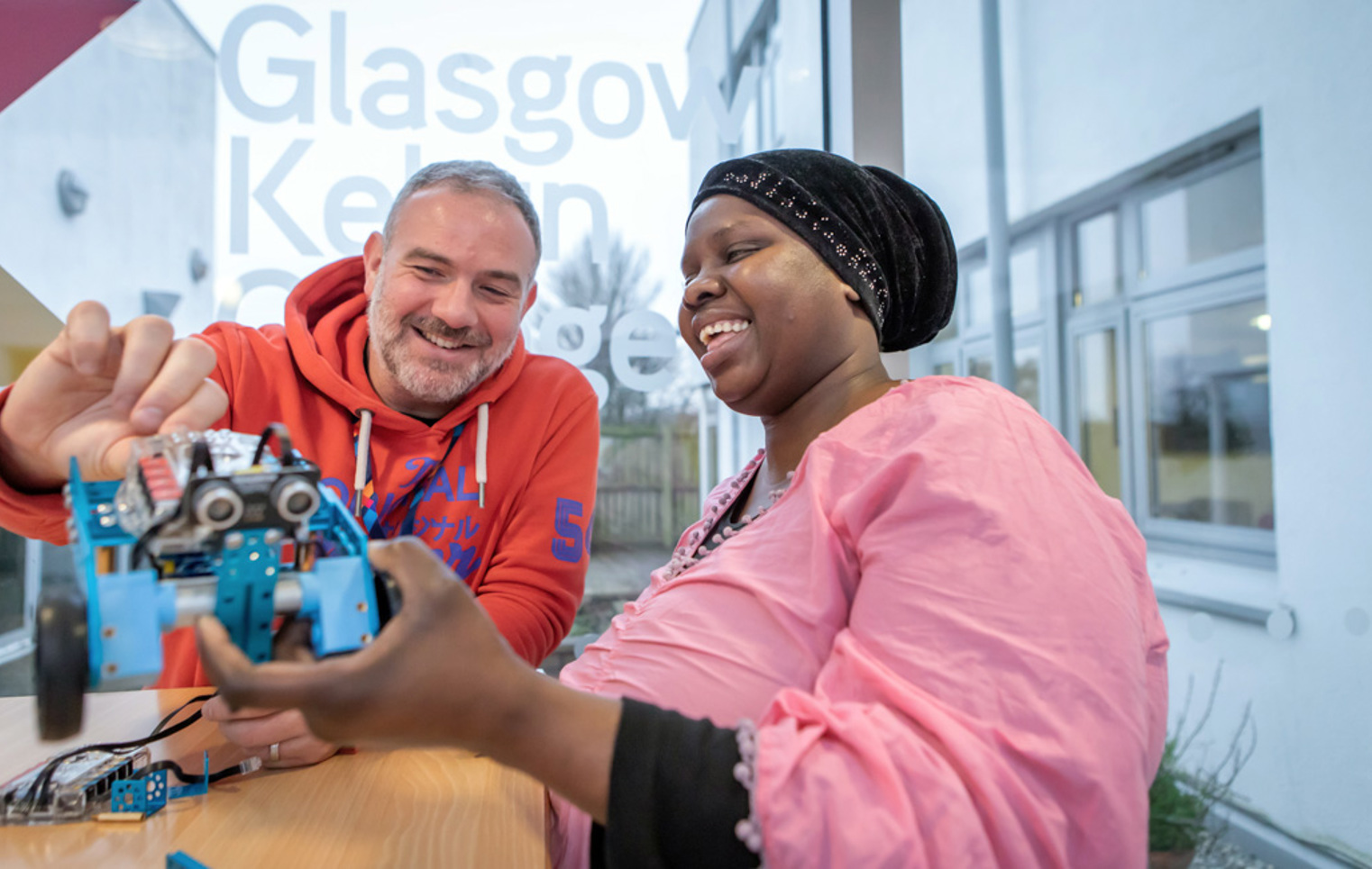 Lecturer in an orange hoodie and a student in a pink top smiling while working together on assembling a blue robotic vehicle near a large window with Glasgow Kelvin College branding