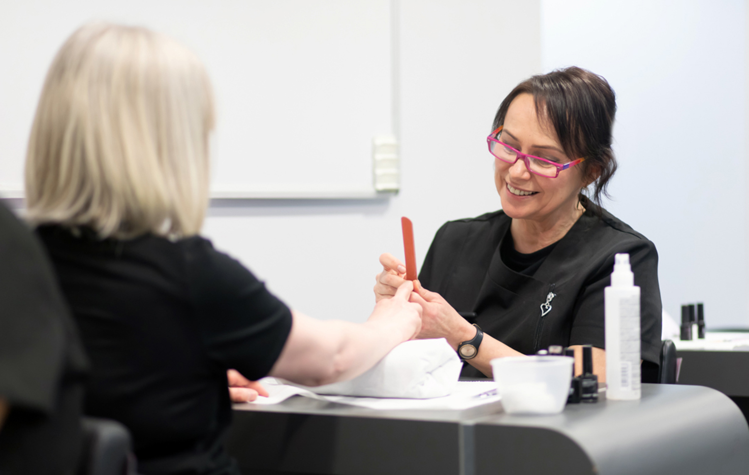 A beauty therapy student provides a manicure to a client, shaping nails with a file while smiling in a professional training setting.