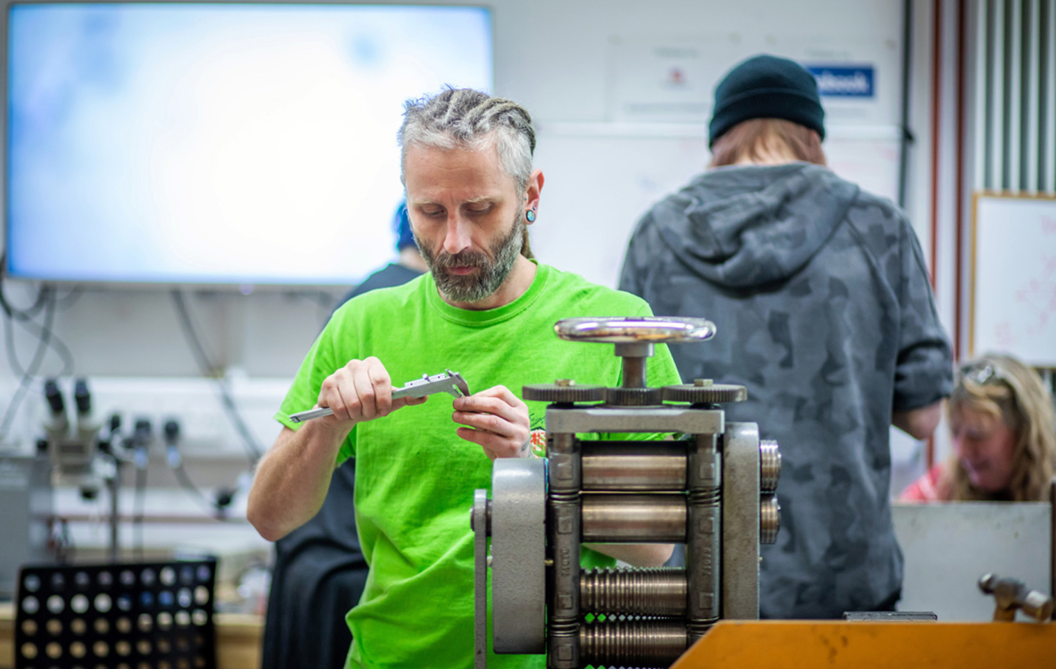 A male craftsman with braided hair and a beard, wearing a bright green shirt, measuring a piece of jewellery with calipers in a workshop filled with equipment.