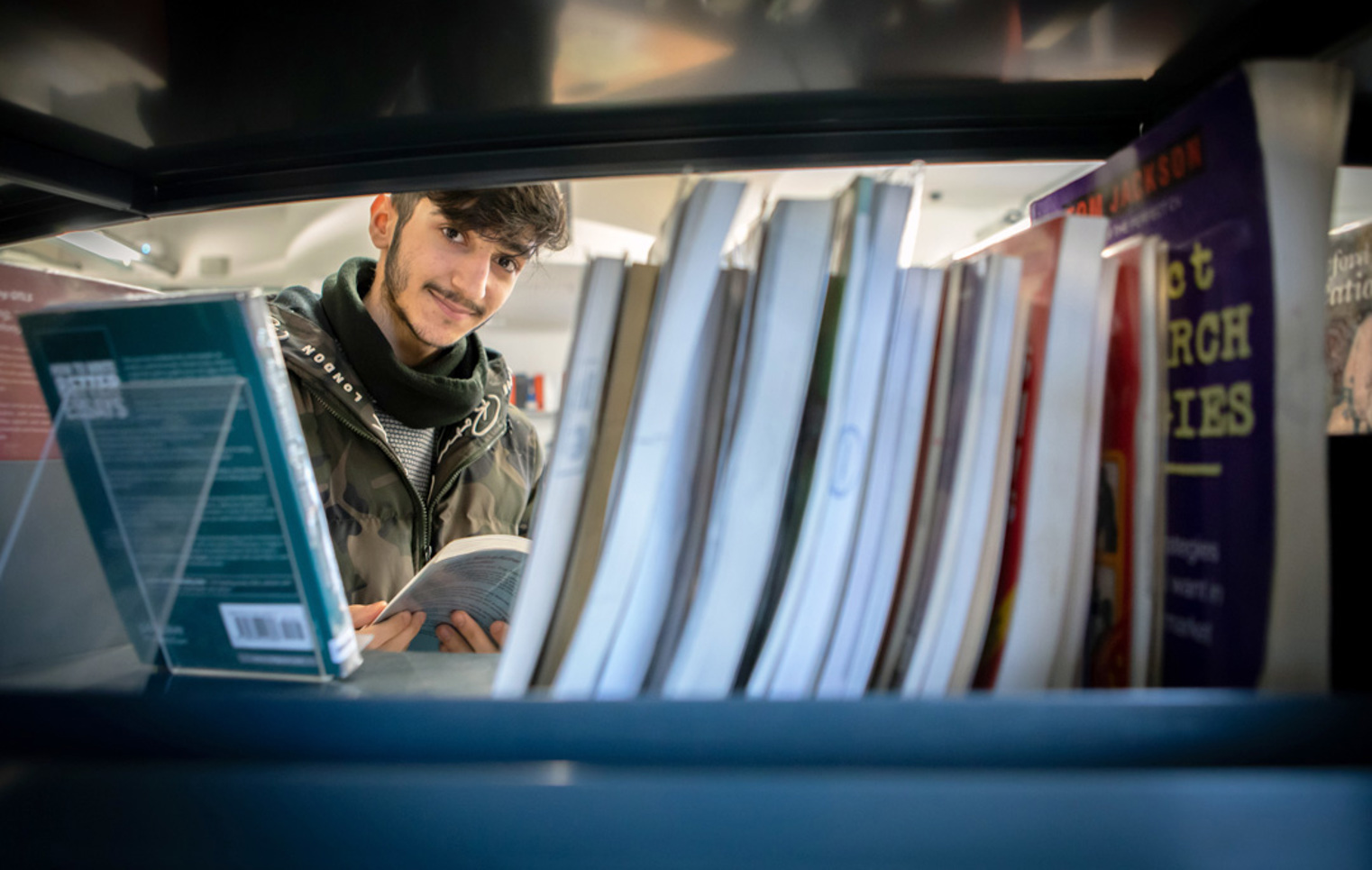 Student browsing books in the East End campus library at Glasgow Kelvin College, viewed through a shelf.