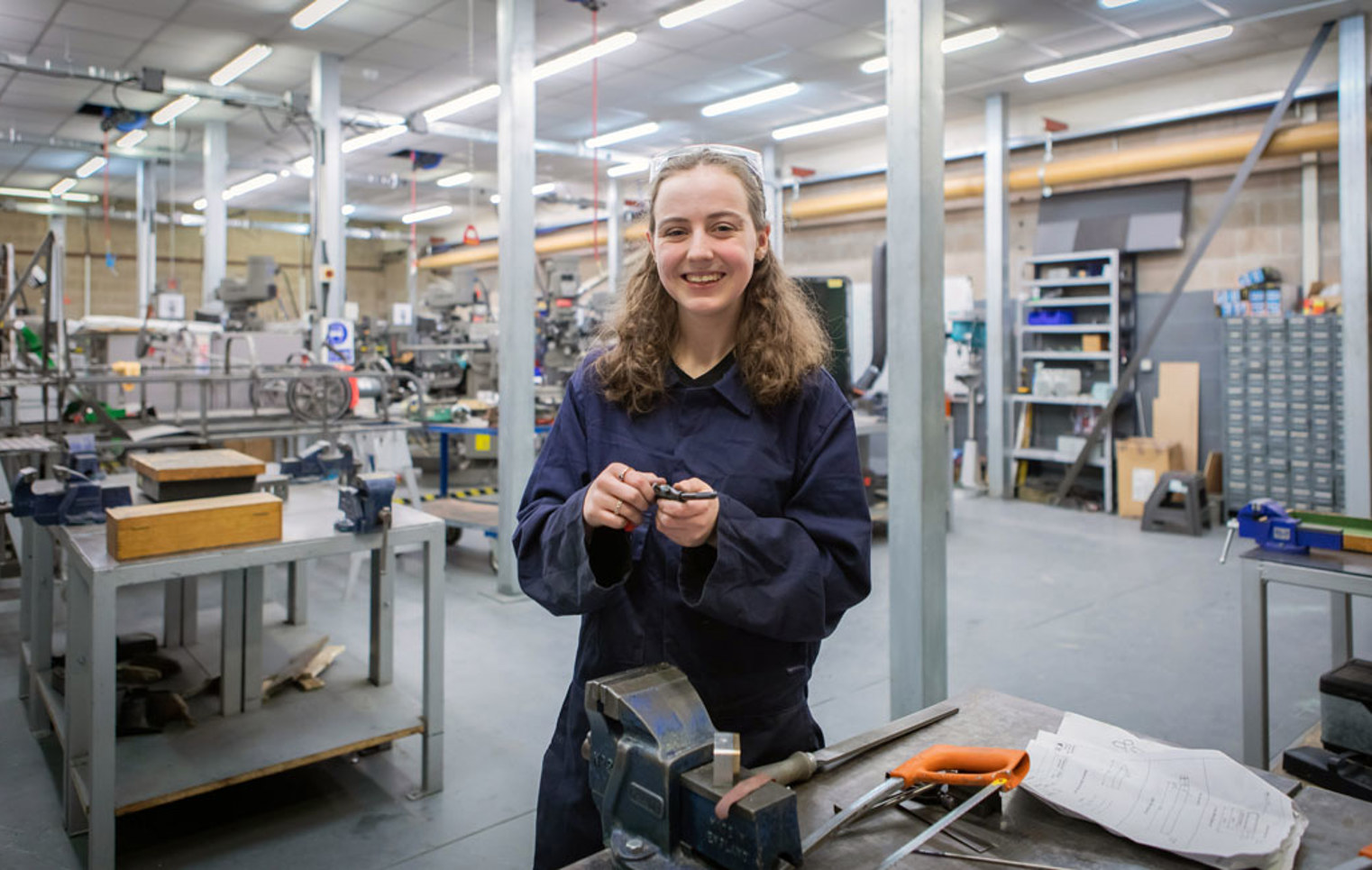 A female mechanical engineering student in navy-blue overalls smiling while working on a small metal component in a busy college workshop.