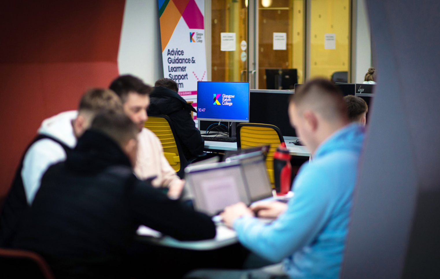 Students working together on laptops in a pod area at Glasgow Kelvin College, with a support services banner and desktop monitors in the background.