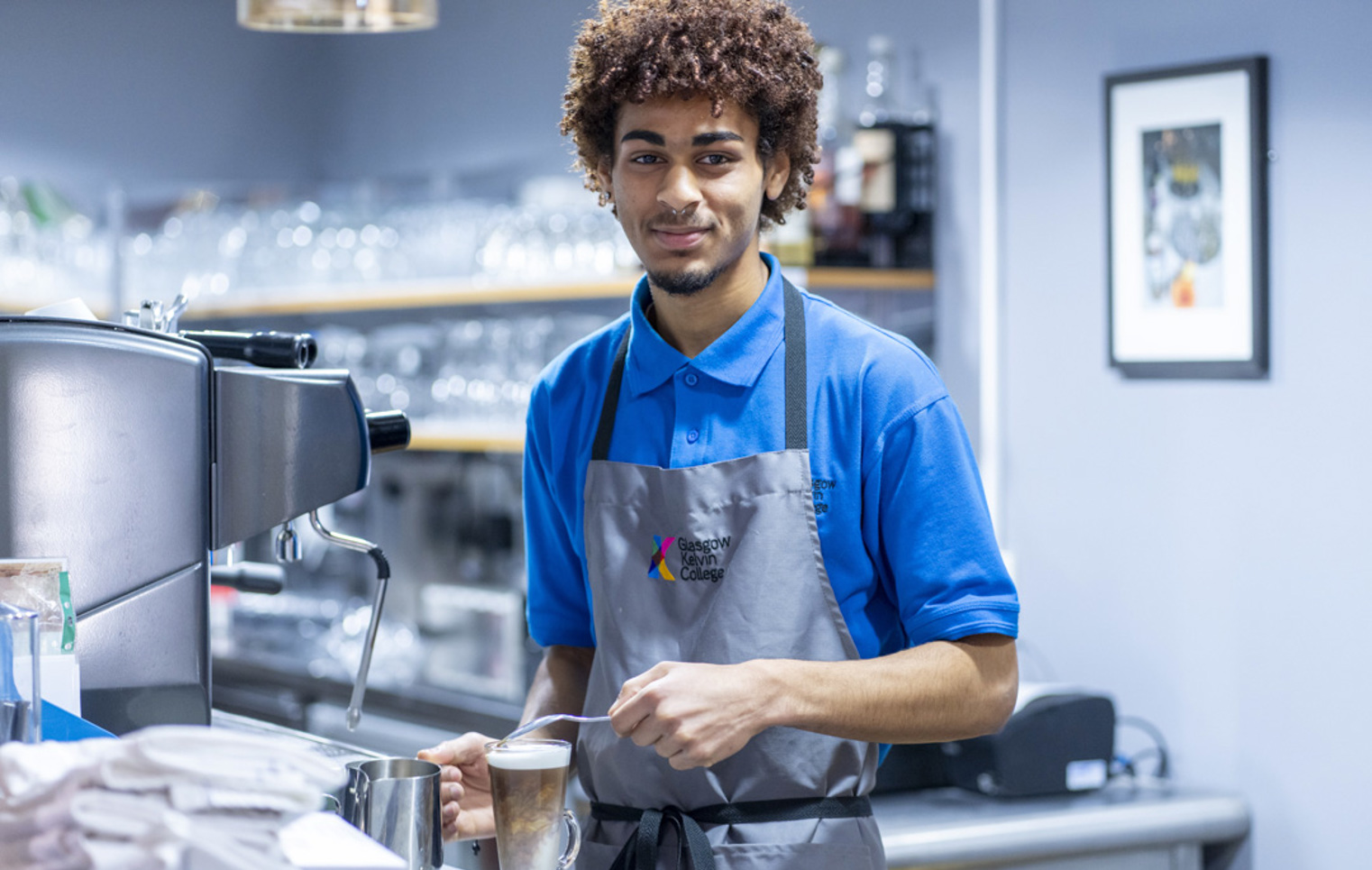 A young male barista in a blue uniform and grey apron, smiling while preparing a latte at a coffee machine in a café setting.