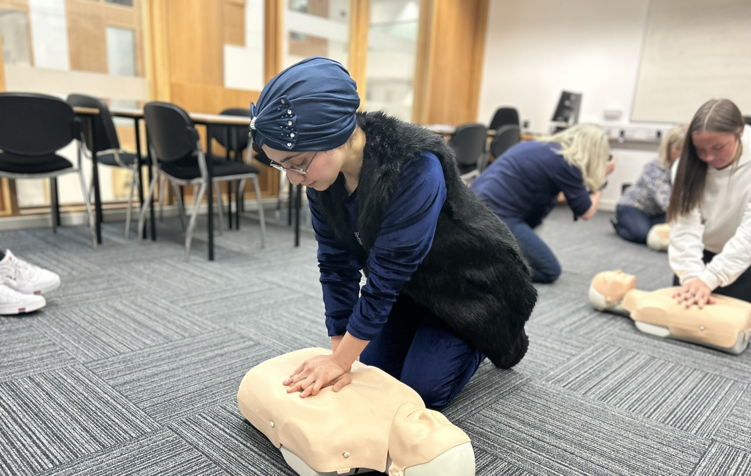 A woman wearing a blue headscarf and glasses practices CPR on a mannequin during a first aid training session in a classroom setting. Other participants are visible in the background.