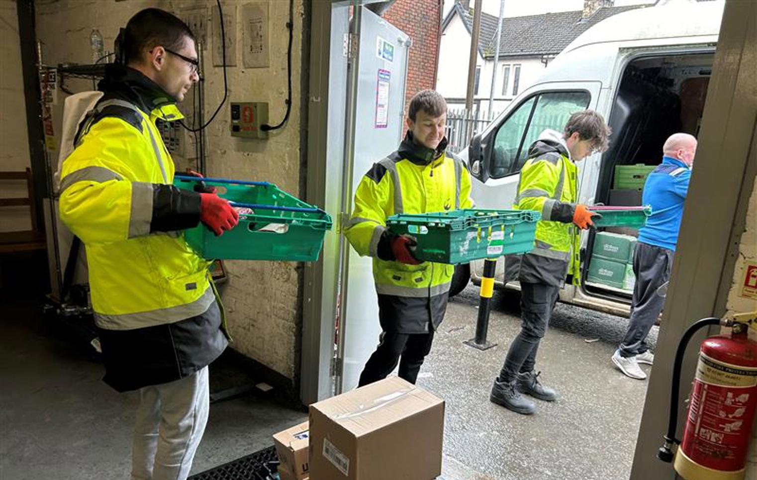 Three young men in high-visibility jackets unloading green crates from a delivery van at a warehouse entrance.