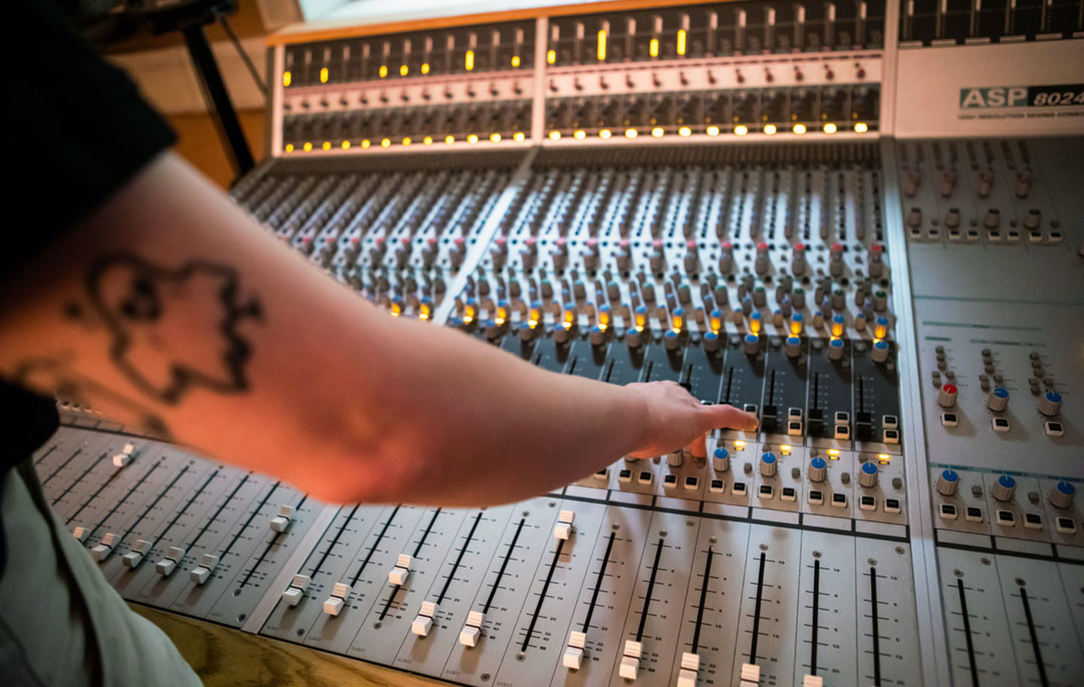 A close-up of a student's arm with a ghost tattoo reaching for controls on a large mixing desk, with glowing indicator lights.