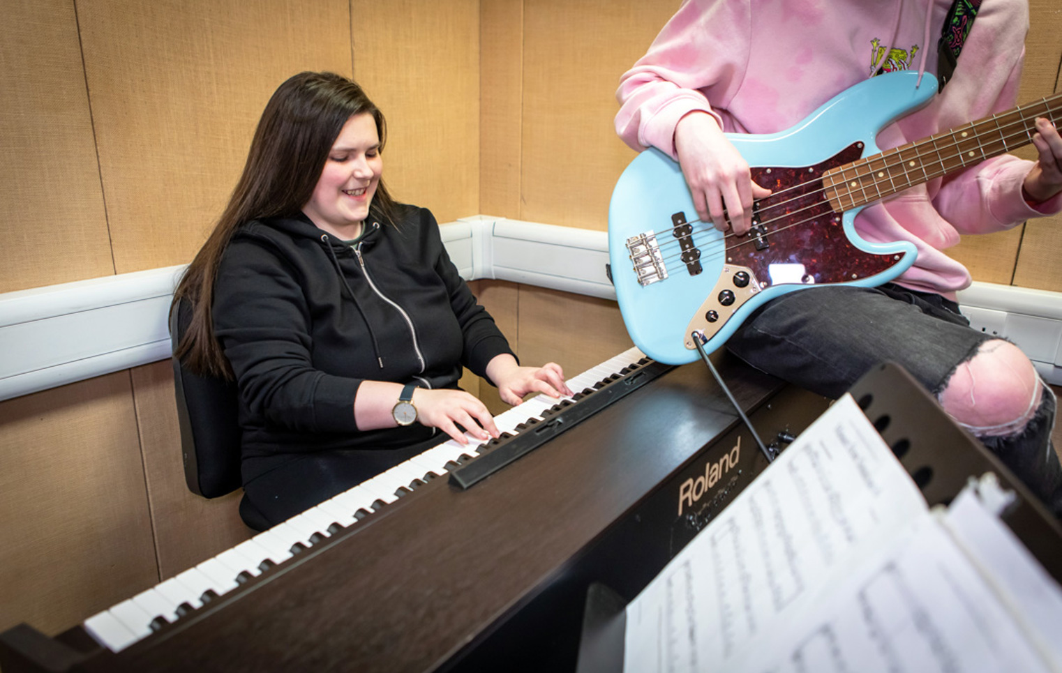 A smiling student in a black hoodie plays a Roland keyboard while another plays a blue bass guitar beside them.