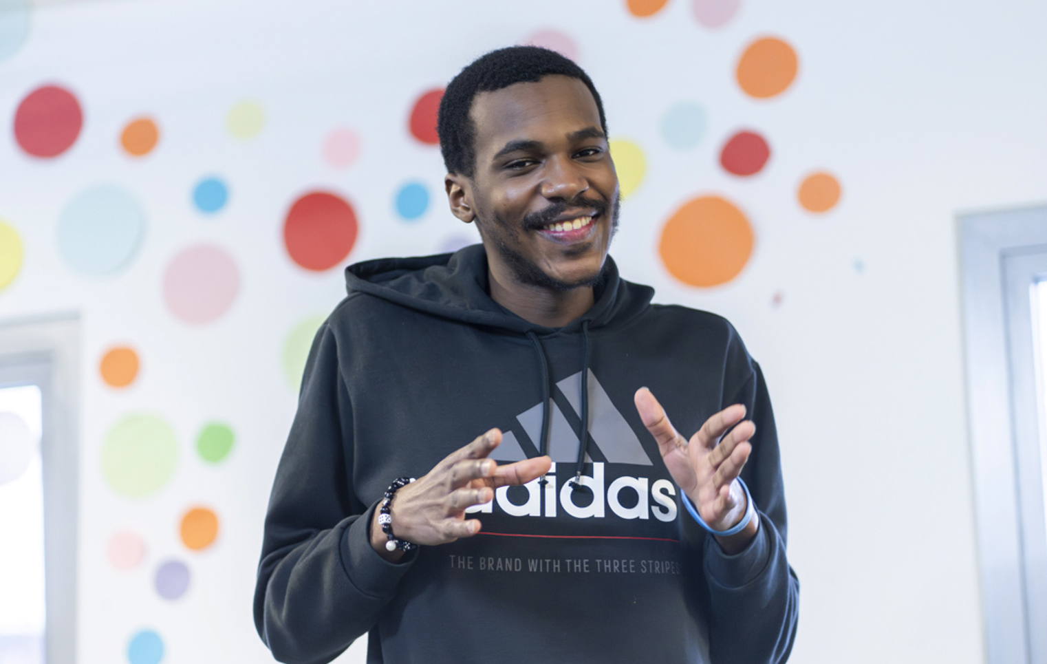 Young man smiling while making expressive hand gestures in front of a colorful polka-dot wall.