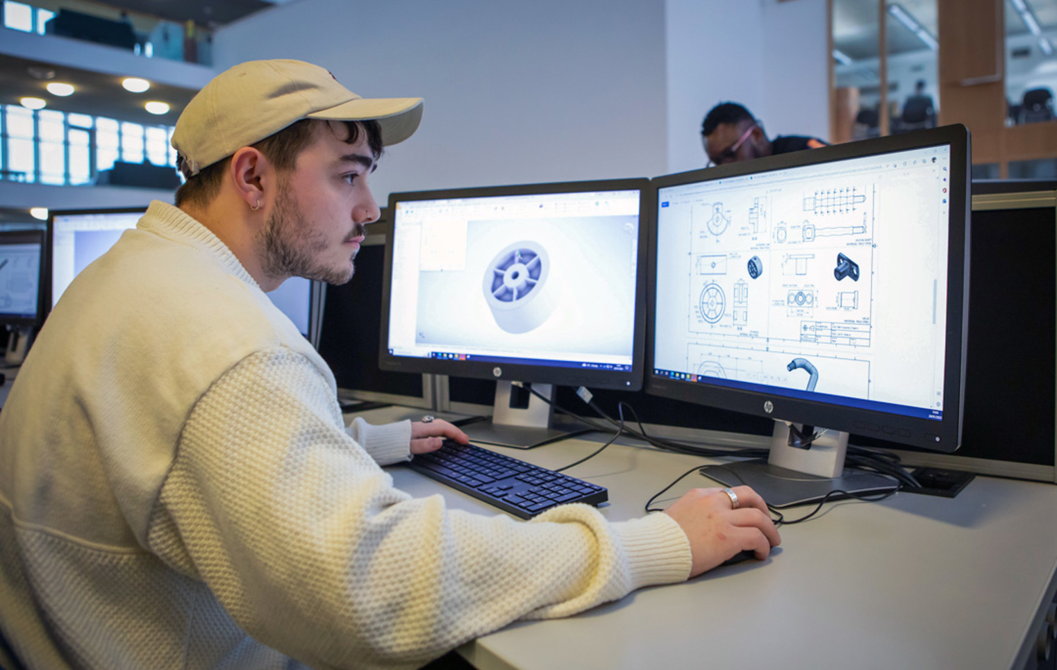A student wearing a white cap working on a dual-monitor CAD setup, designing a mechanical component on one screen and a technical drawing on the other.