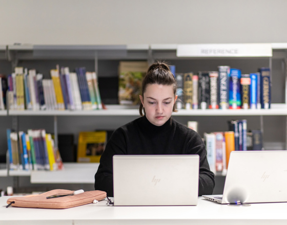 Student working on a laptop in the library at Glasgow Kelvin College, seated at a desk with bookshelves in the background.