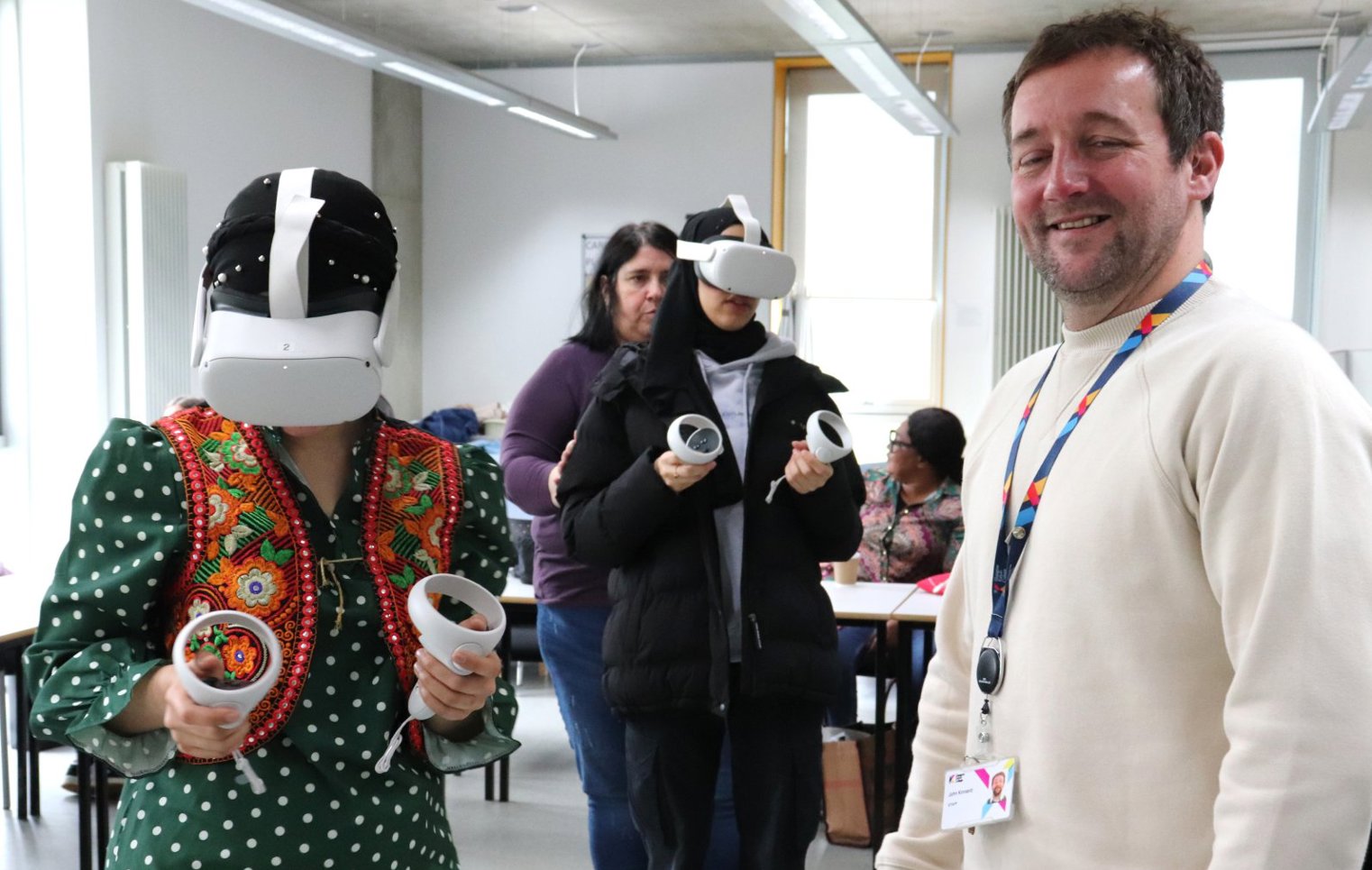 A smiling instructor stands in the classroom while a student wearing a VR headset and traditional attire engages in a virtual training session, with other students participating in the background.