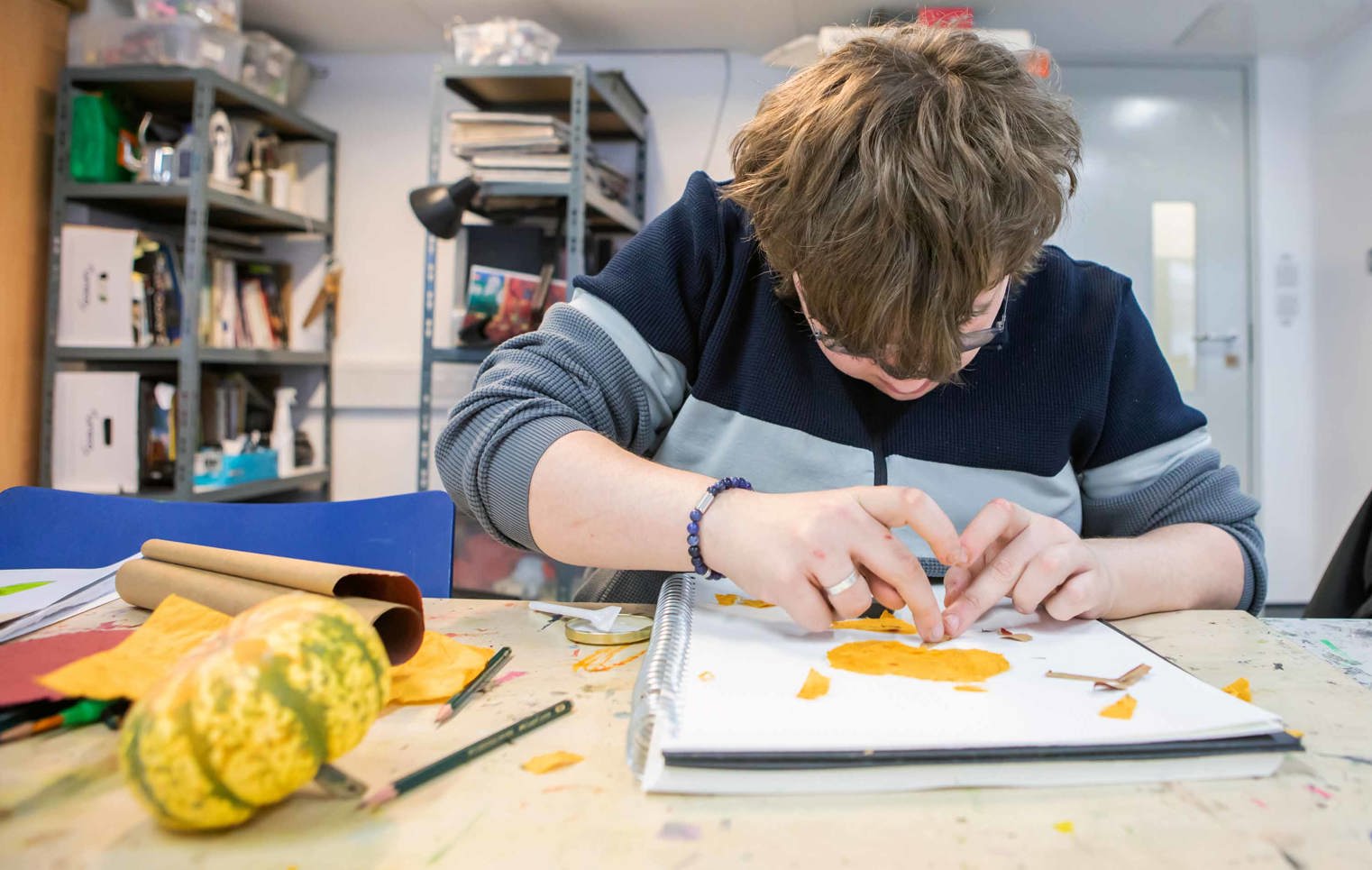 Student working on an art project, using orange paper to create a design in a sketchbook, surrounded by art supplies.