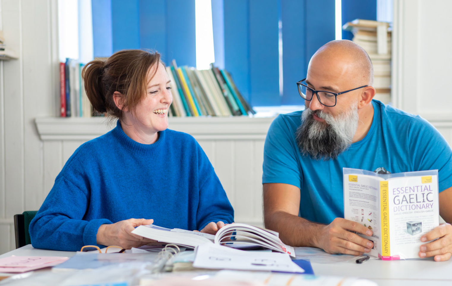 Two students in a Gaelic language class laughing while studying, with a Gaelic-English dictionary open on the desk.