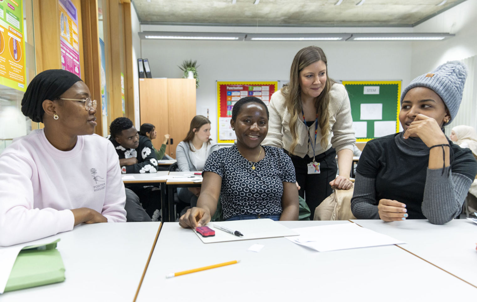 A classroom scene where three female students are smiling and engaging in discussion. A teacher in a beige cardigan leans over the table to interact with them, while other students work in the background.