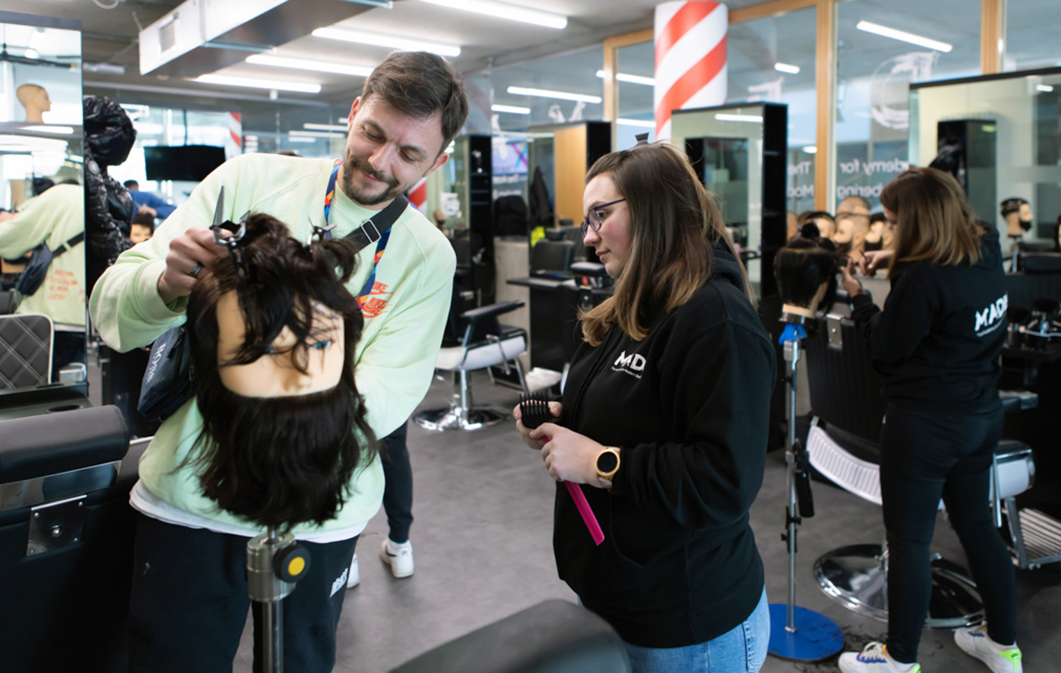 A male instructor in a light green sweatshirt assisting a female student with a mannequin head in a barbering classroom.