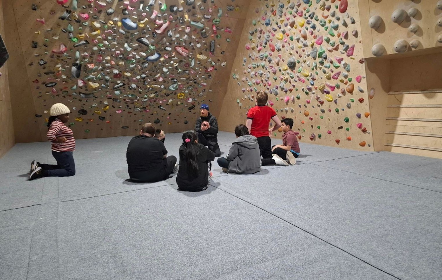 Supported Learning students sitting with an instructor at an indoor climbing centre, listening during a group briefing beneath a bouldering wall.