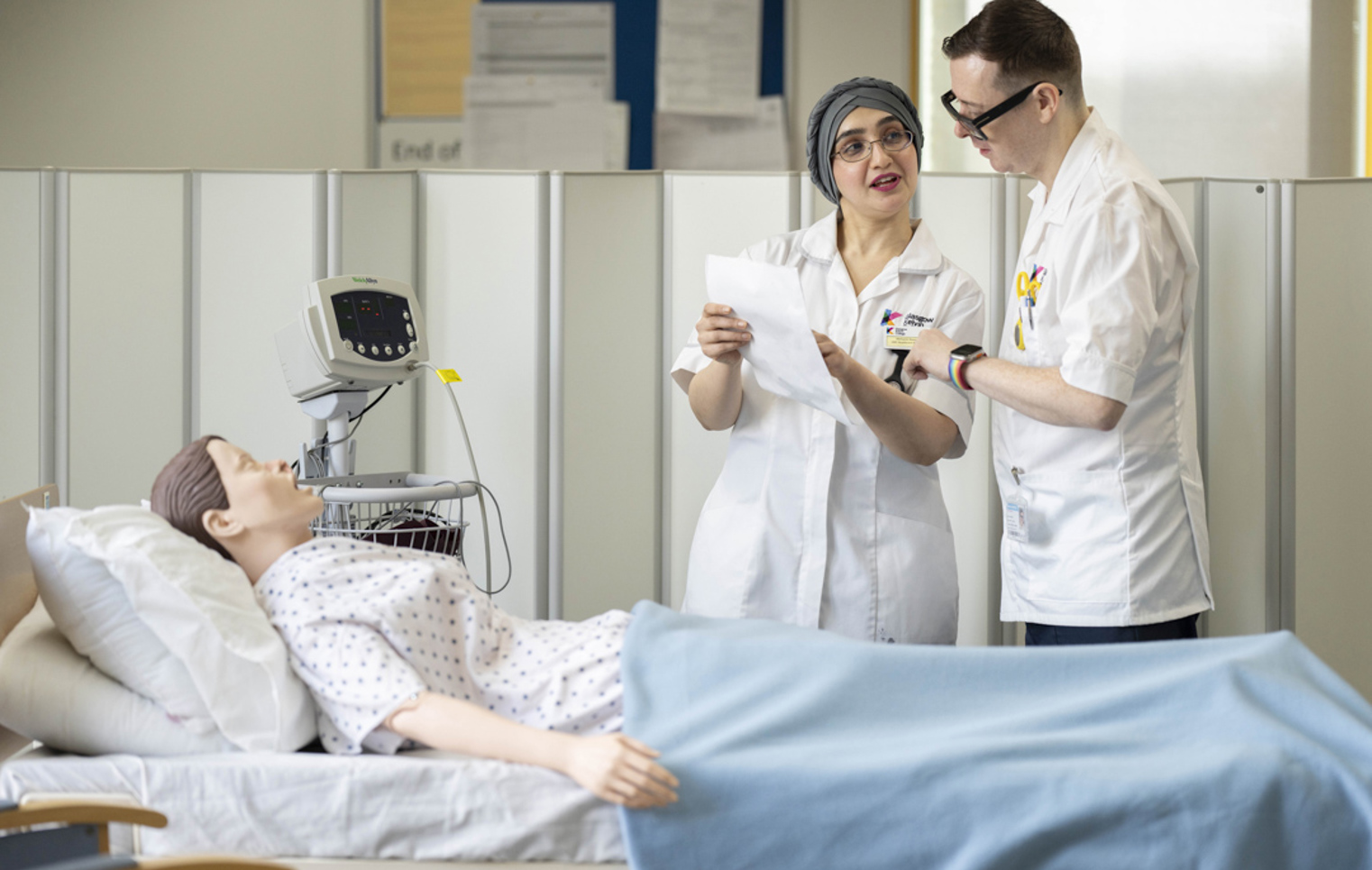 Two care students discuss notes at a hospital bed during a training exercise