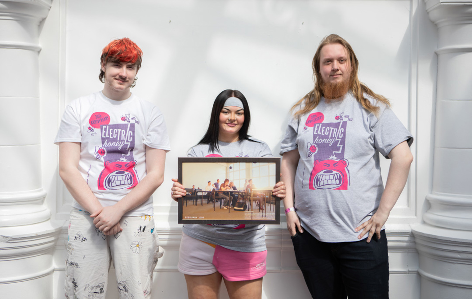 Three students wearing Electric Honey t-shirts standing in front of a white wall, holding a framed classroom photo from 2008.