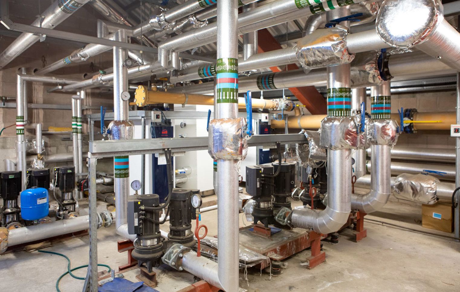A network of insulated industrial pipes, pumps, and control valves in a large building services training facility