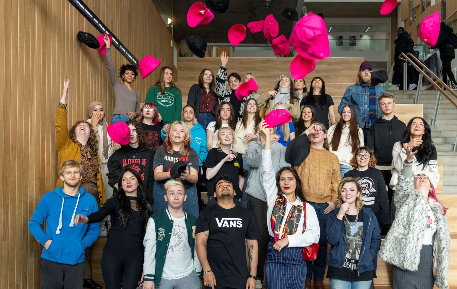Large group of students gathered on indoor wooden steps, smiling and tossing bright pink and black hats into the air in celebration.