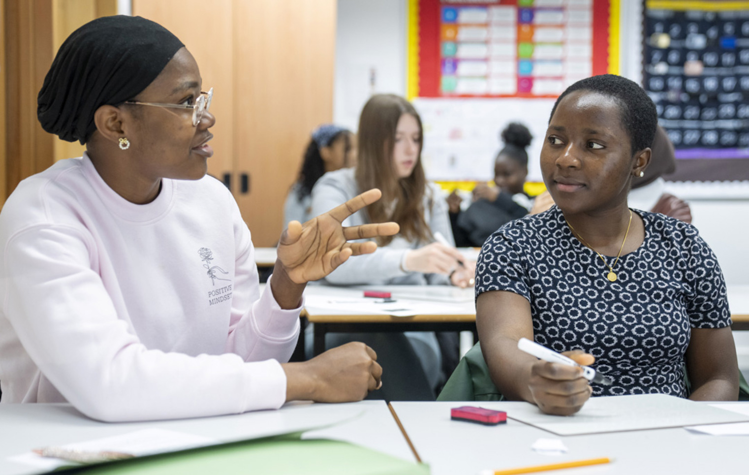 Two female students sit at a desk in a classroom. One student in a pink sweatshirt and a black headscarf is gesturing as she speaks, while the other in a patterned dress listens attentively with a pencil in hand