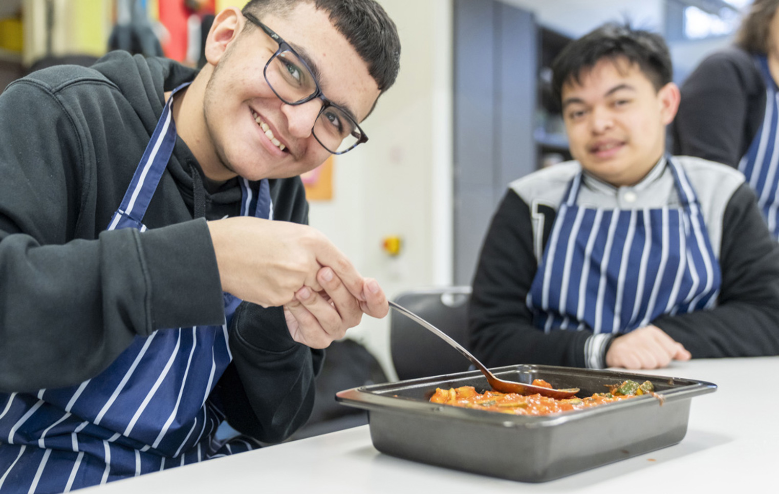Two young men smiling and preparing a tray of food, wearing striped aprons in a kitchen setting.