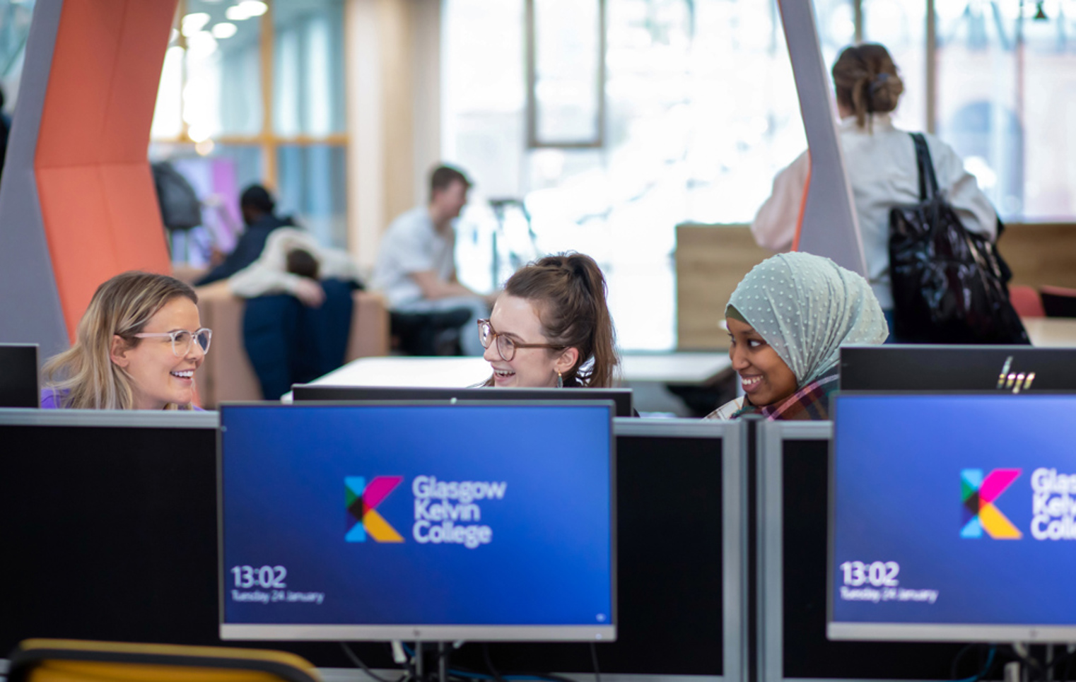 Three female students smiling and chatting while using desktop computers in the library area at Glasgow Kelvin College’s Springburn Campus.