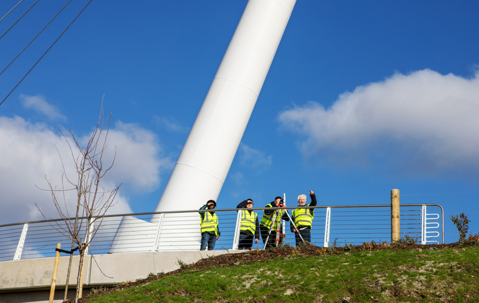 Wide-angle view of the Stockingfield Bridge with a group of students and lecturers in high-visibility vests conducting a surveying exercise on the platform beneath a bright blue sky.
