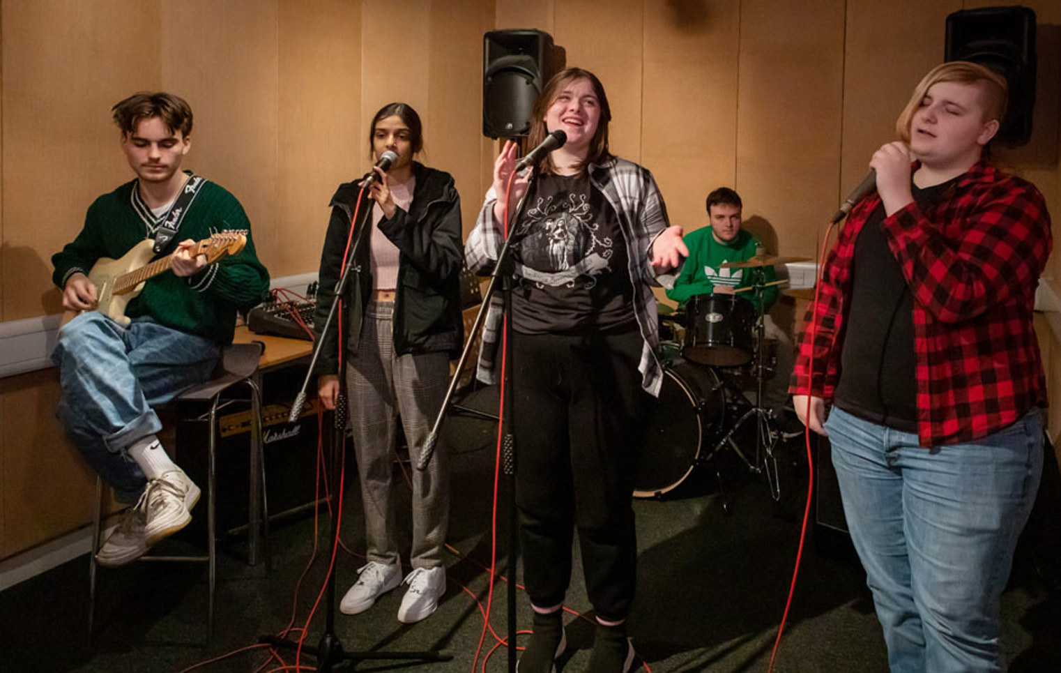 A group of five students rehearsing, featuring a guitarist, two vocalists, a bassist, and a drummer in a music studio.