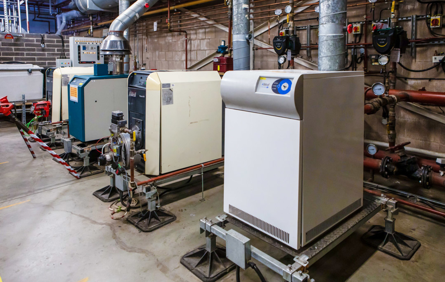 A row of industrial boilers in a building services training facility, connected to ventilation ducts and a complex piping system.