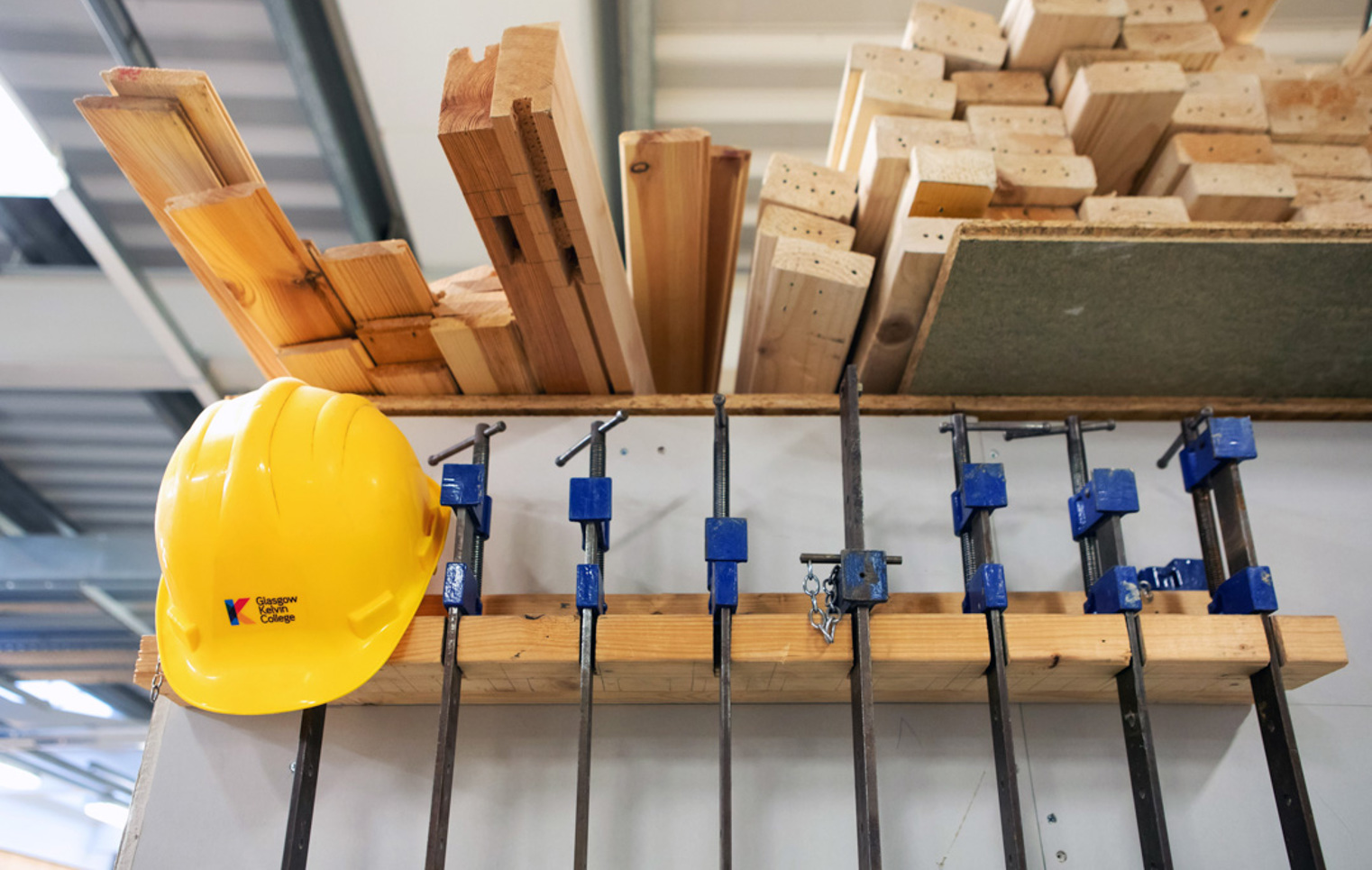Woodworking equipment including clamps, timber planks, and a bright yellow safety helmet neatly arranged on a storage rack in a joinery workshop.