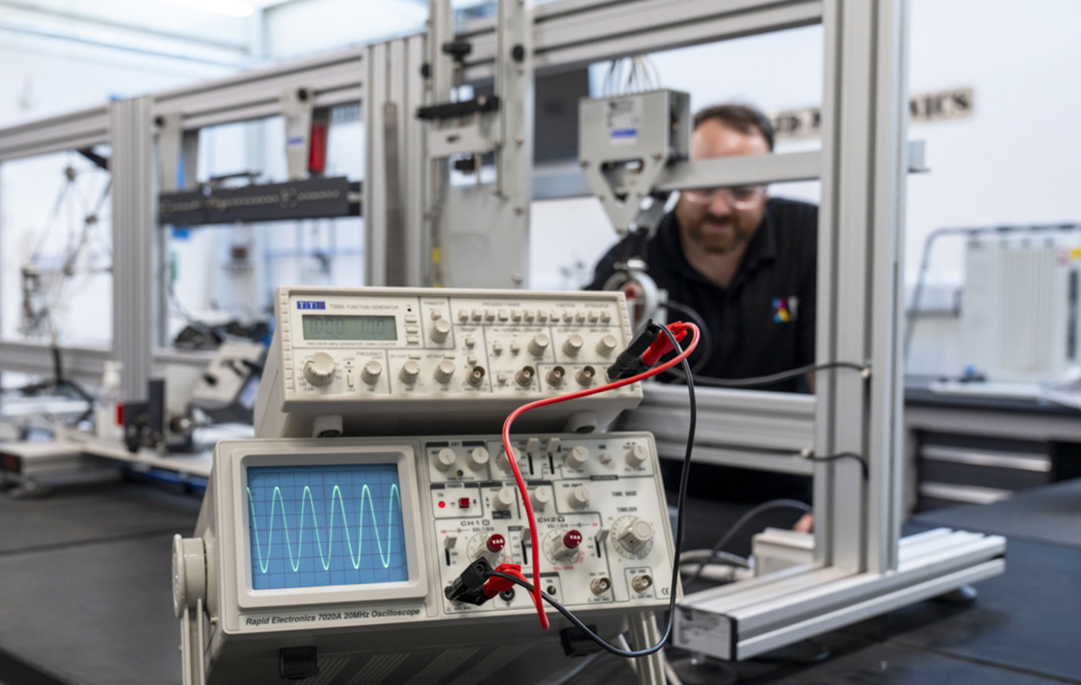 Close-up of an oscilloscope displaying a waveform, connected to a function generator in a laboratory, with a lecturer working in the background.
