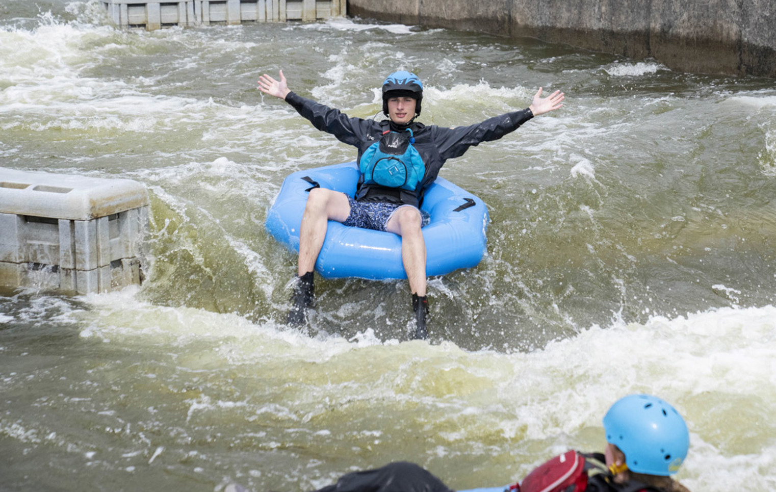 A student with arms outstretched riding a blue inflatable ring on a whitewater course.