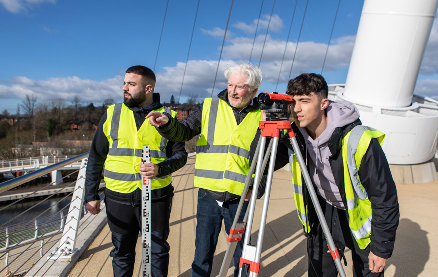 Three individuals wearing high-visibility vests conducting a land surveying exercise on a bridge. One is looking through a theodolite, while another holds a measuring staff, and a lecturer observes under a bright blue sky.