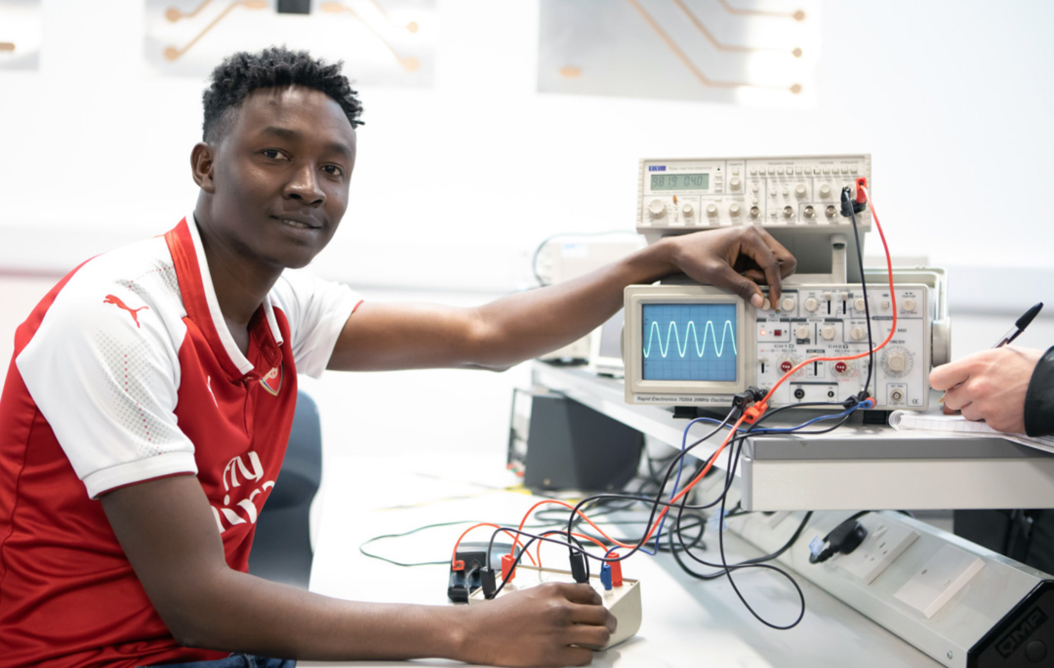 A student in a red football jersey adjusting an oscilloscope in an electronics lab, looking at the camera with a confident expression.