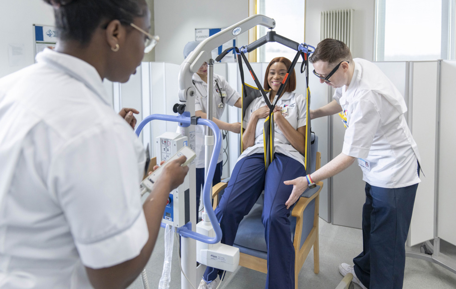 Care students practice using a patient hoist with a seated classmate in a simulated care setting.