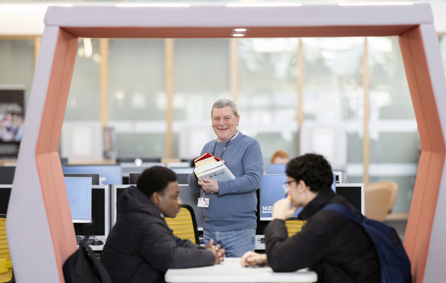 Smiling staff member holding books and standing beside two students in a Glasgow Kelvin College library study pod.