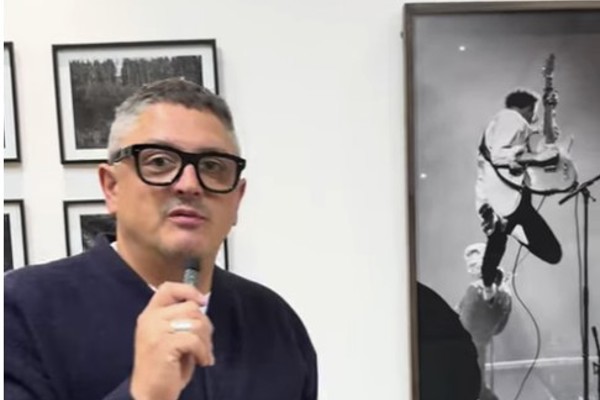 Photographer Mark Cameron speaking at an exhibition, standing in front of a wall of framed black and white portraits, including an action shot of a guitarist.