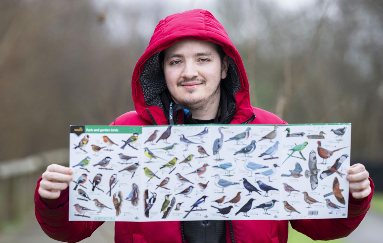 Smiling man in a red jacket holding a bird identification poster with various bird species displayed.