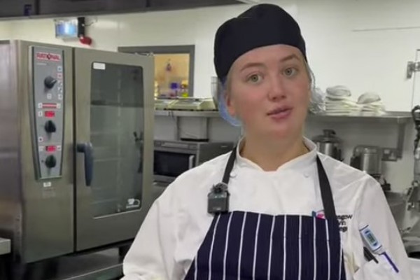 A cookery student in a striped apron and black chef's hat uses a blowtorch to char sweetcorn in a commercial kitchen.